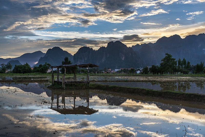 Water_reflection_of_a_wooden_hut_with_colorful_sky_and_karst_mountains_in_a_paddy_field_at_sunset_Vang_Vieng_Laos